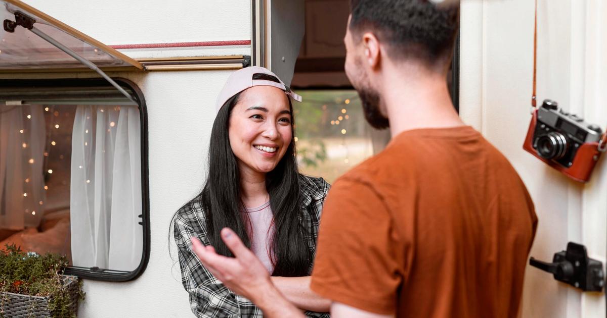 Young couple conversing outside an RV with open window, smiling and engaged in friendly discussion near the camper entrance.
