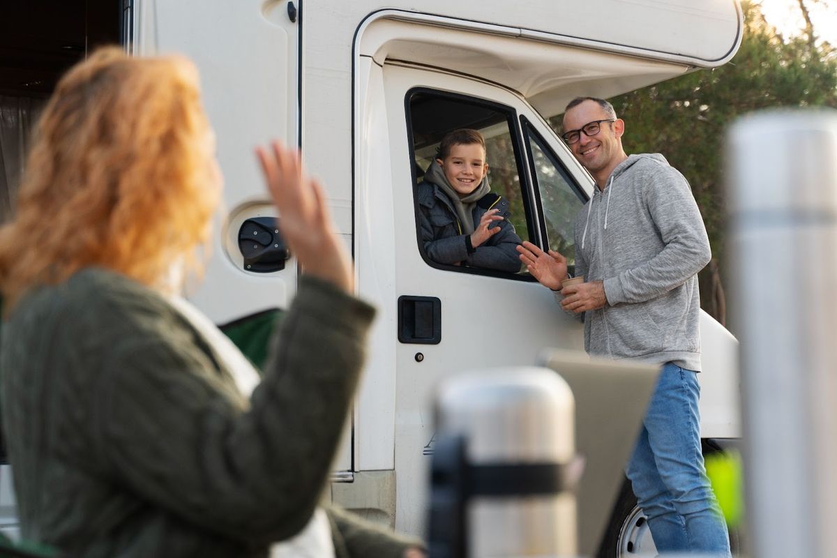 Family waving from RV doorway during outdoor visit, man and boy smiling inside camper while woman waves goodbye outside.