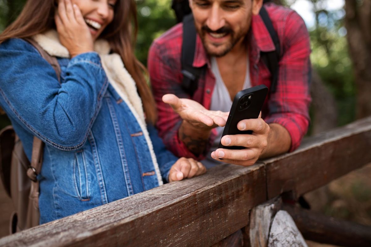 Couple outdoors by wooden fence viewing smartphone together, both smiling in natural setting with trees in background.