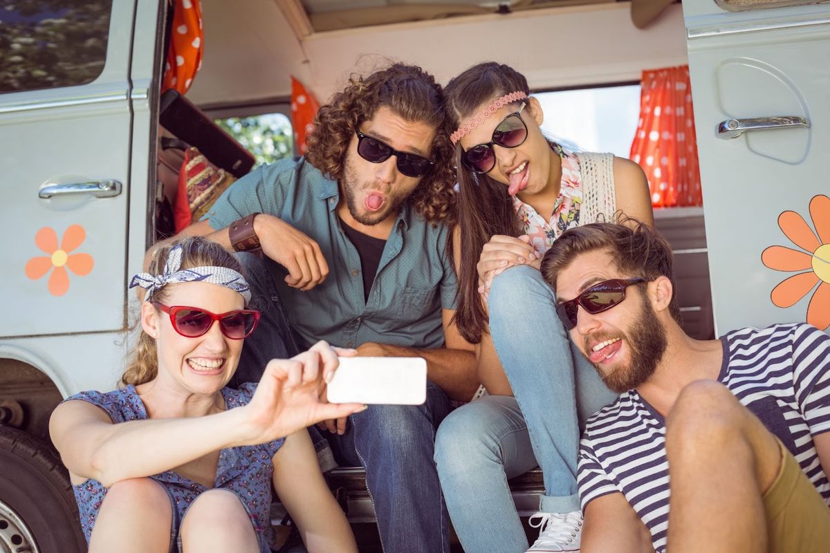Five friends taking a selfie inside a retro camper van decorated with orange flower decals, all wearing sunglasses and making playful faces.