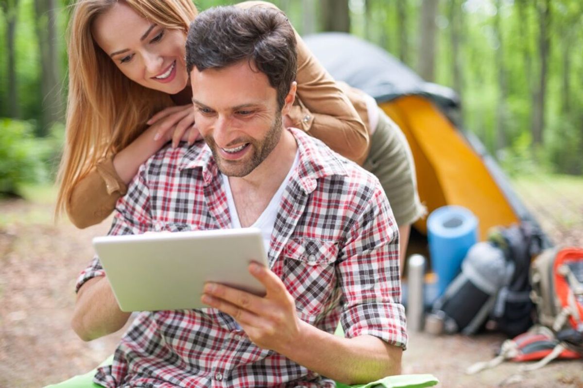 Smiling couple at a forest campsite reviewing information on a tablet, with a tent and camping gear behind them