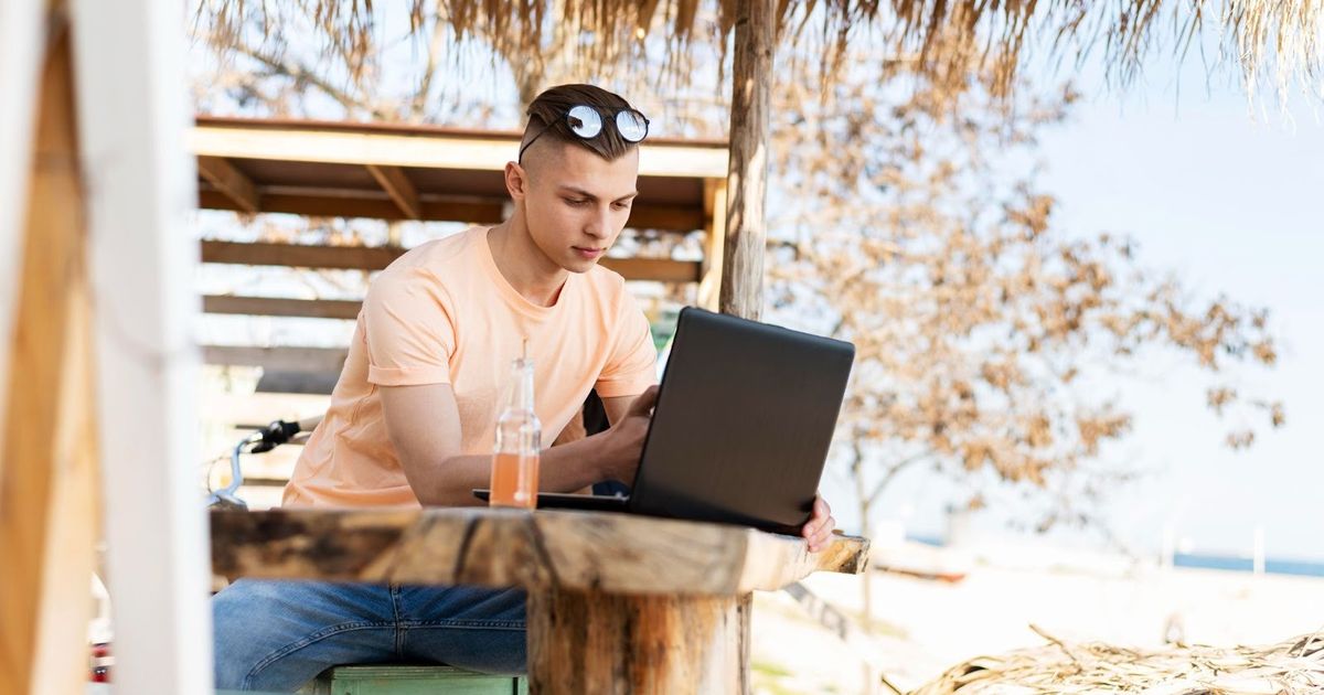 Young man working on laptop at wooden table under thatched roof structure, with glass of juice beside him, sunglasses on head.