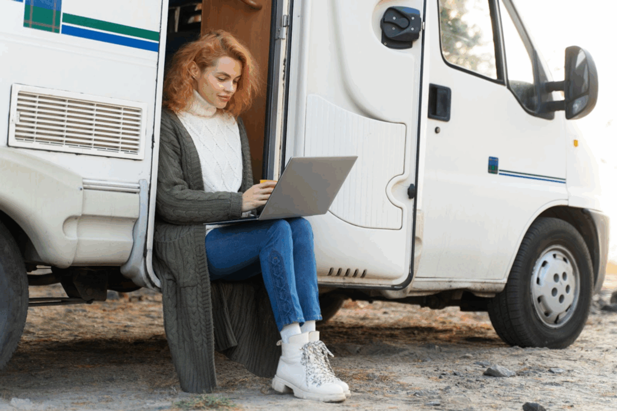 Woman with red hair sitting in RV doorway working on laptop, representing remote work and digital nomad lifestyle at campgrounds.