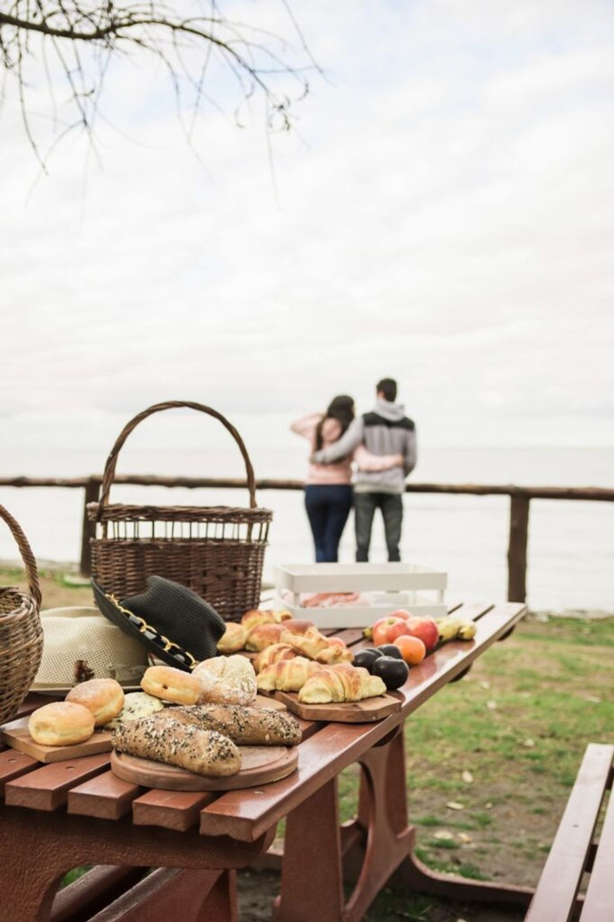 Picnic spread with fresh bread, croissants, and fruit on a wooden table with wicker baskets, couple embracing by waterside railing in background.