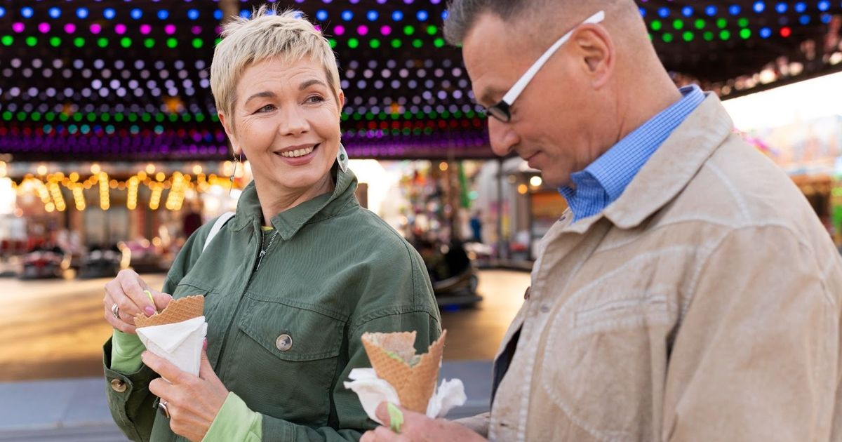 Two people enjoying ice cream cones at a colorful illuminated fair or amusement park with rides visible in the background.
