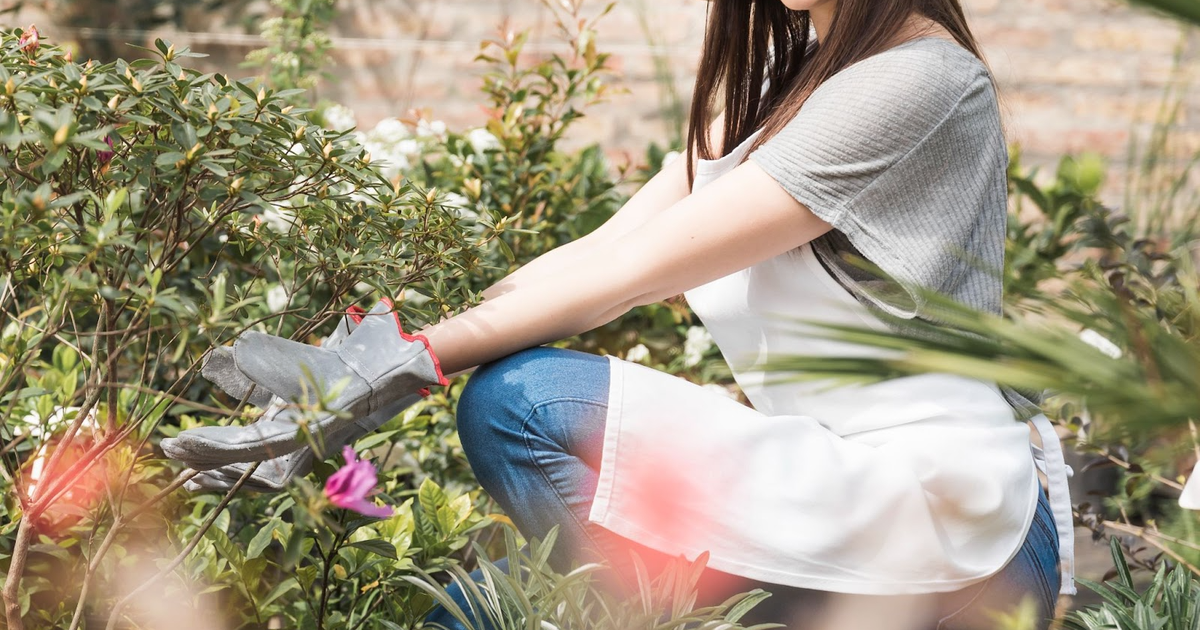 Woman in white sun hat tending to garden plants against brick wall, wearing gardening gloves and smiling at camera.