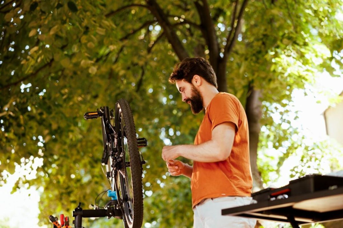 Man in orange shirt repairing bicycle outdoors under leafy trees in a park setting.