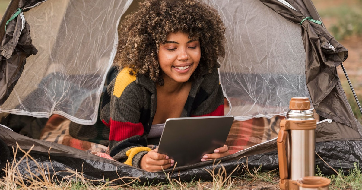 Young woman with curly hair smiling while using a tablet inside a tent during rainfall, with a thermos nearby.