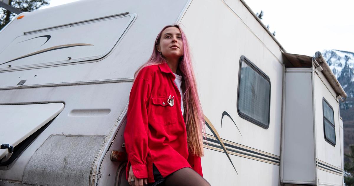 Woman with pink hair in red jacket standing on RV steps, mountains and forest visible in background.