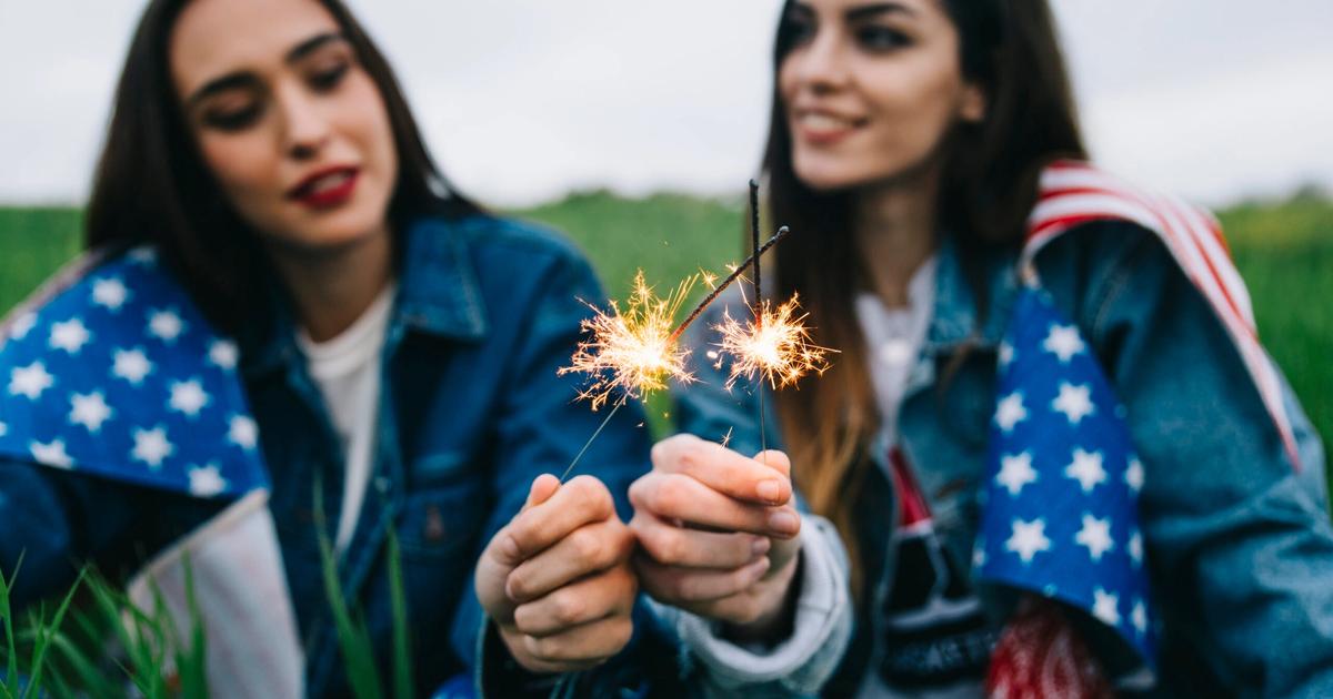 Two women holding lit sparklers outdoors in a grassy field, wearing patriotic-themed jackets with stars and stripes.