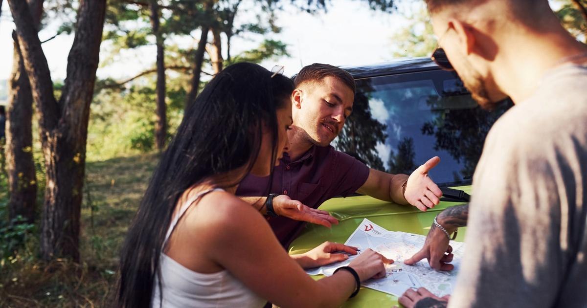 Three people gathered around a vehicle hood studying a map together in a forested campground setting during daytime.