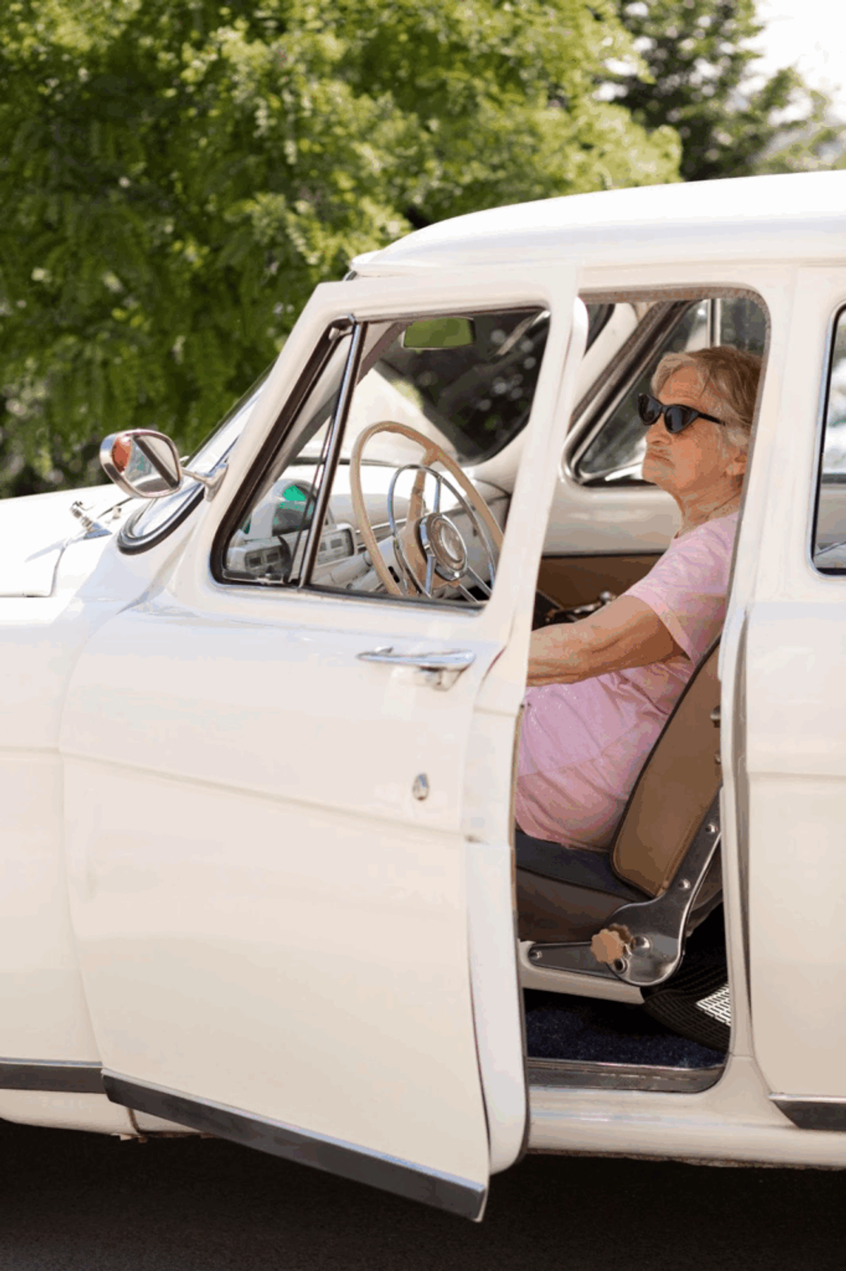 Man in sunglasses and pink shirt sitting in driver's seat of vintage white pickup truck with door open, green trees in background.