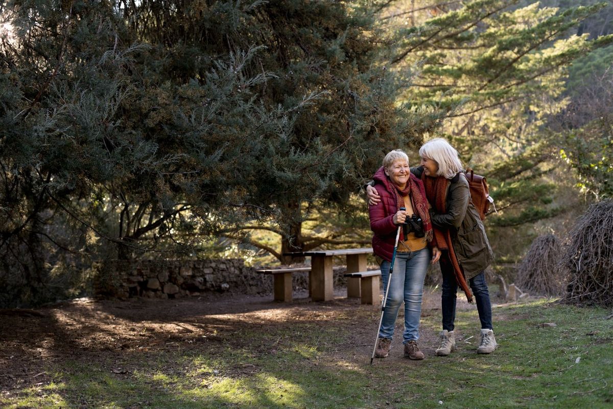Two older adults hiking together on a shaded forest trail near a picnic table, both smiling and enjoying outdoor recreation.