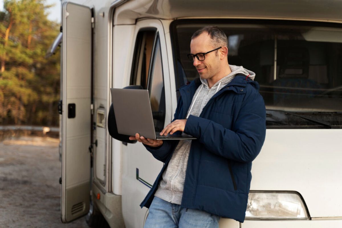 Man with glasses using laptop while standing in front of open door of white RV, wearing dark jacket and gray hoodie.