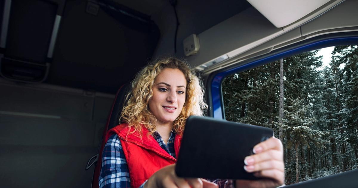 Woman with curly blonde hair in red vest using tablet inside RV, forest visible through window behind her.