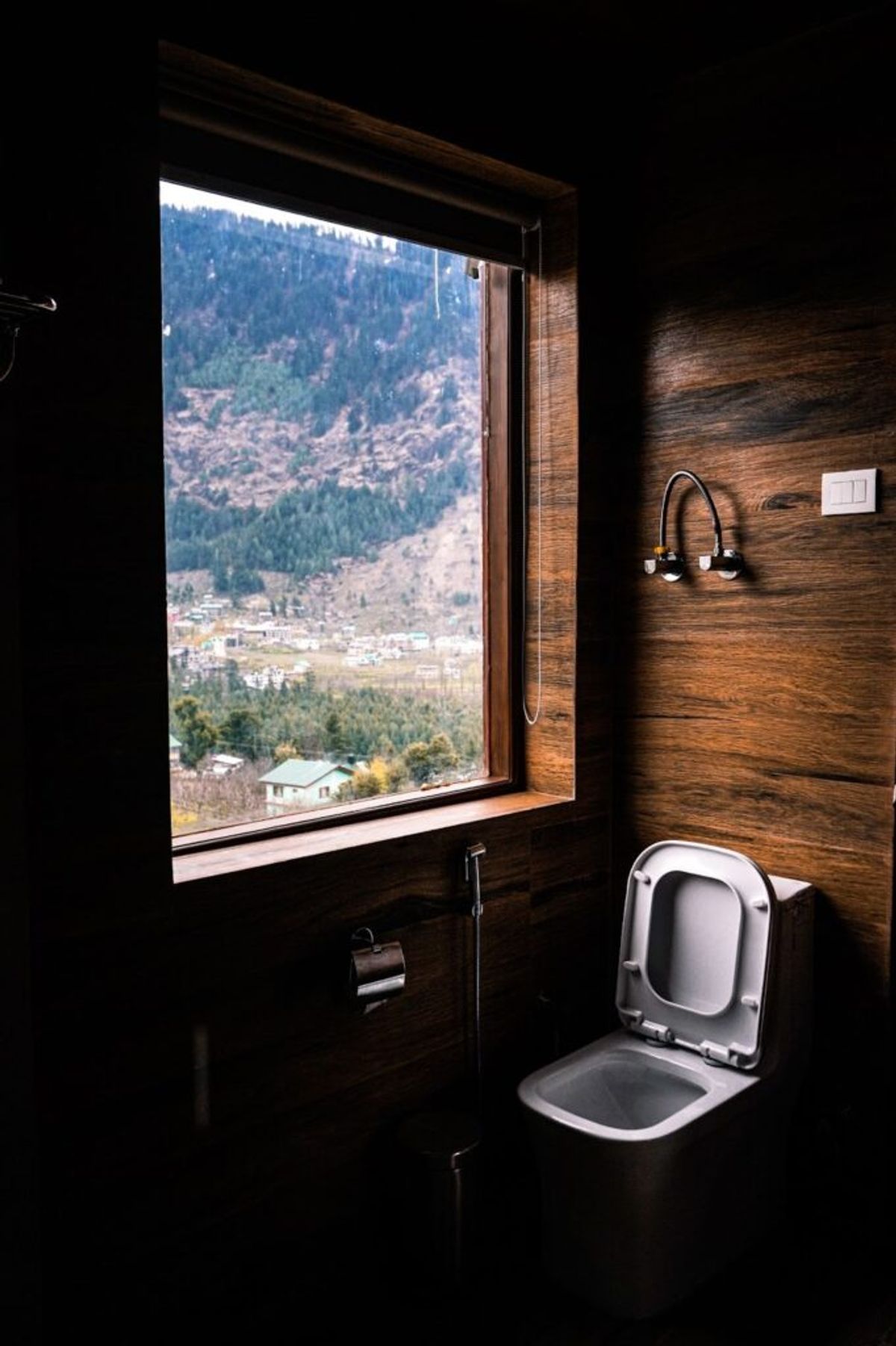 Modern bathroom with wooden walls and an open window overlooking a forested mountain valley with scattered buildings below.