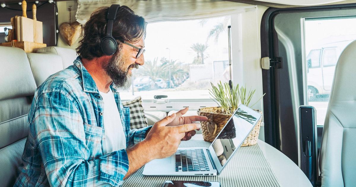 Man wearing headphones working on laptop at a table inside an RV, with palm trees visible through the window.
