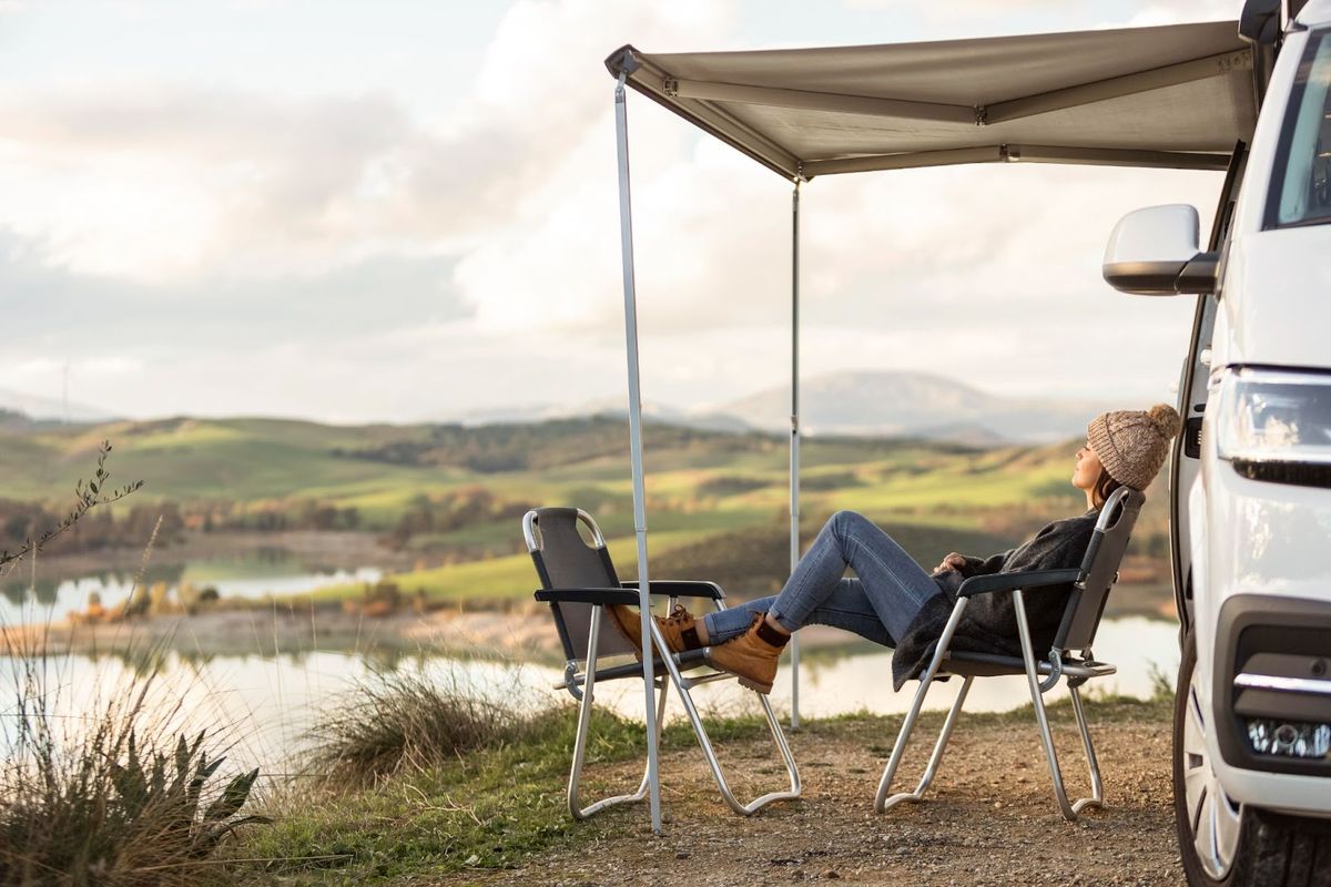 Person relaxing under awning beside campervan with scenic valley landscape, river, and mountains visible in background.