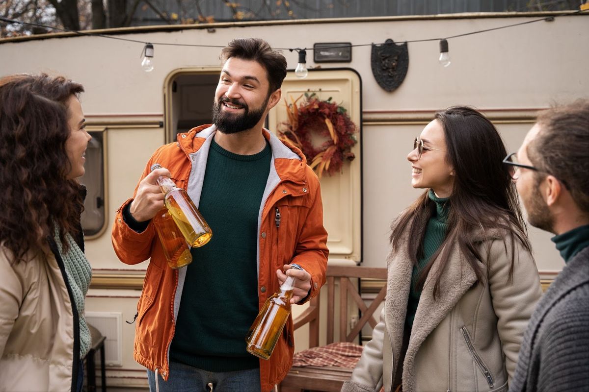 Group of friends socializing outside an RV with bottles of cider, enjoying a festive gathering near a decorated trailer with holiday wreath.
