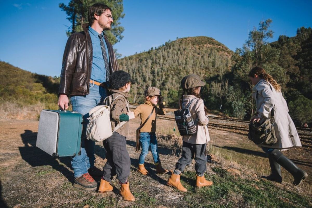 Man and children with luggage and backpacks standing on railroad tracks in forested mountains, preparing for outdoor adventure.
