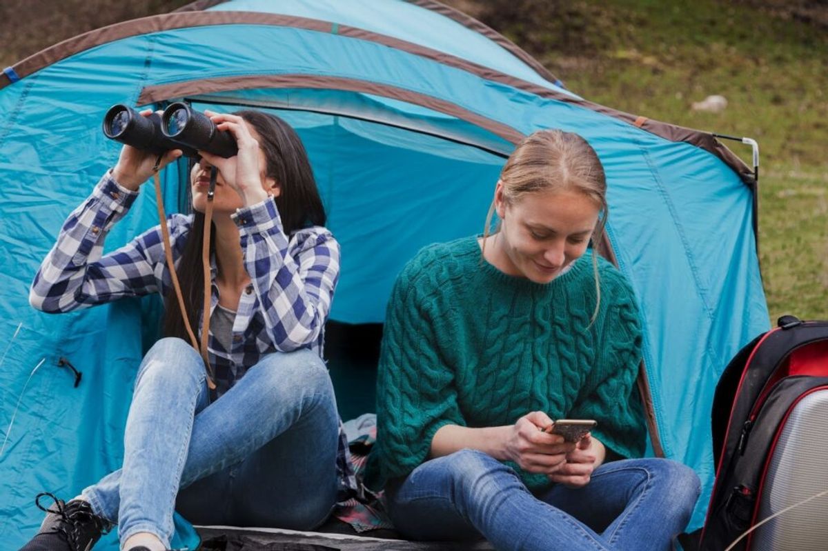 Two young women inside a blue camping tent, one using binoculars to observe wildlife while the other checks a mobile device.