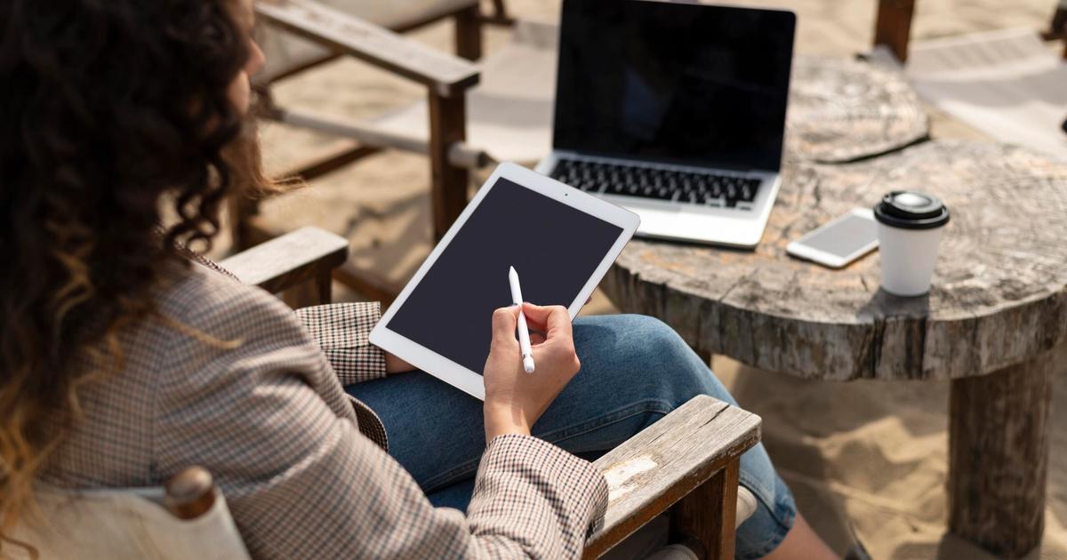 Woman working on tablet with stylus at outdoor wooden table, laptop and coffee nearby on sandy beach setting.