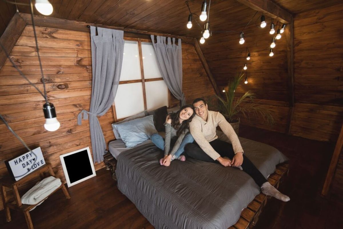 Couple smiling on bed in cozy wooden cabin with string lights, gray curtains, and "Happy Day Love" sign on wall.