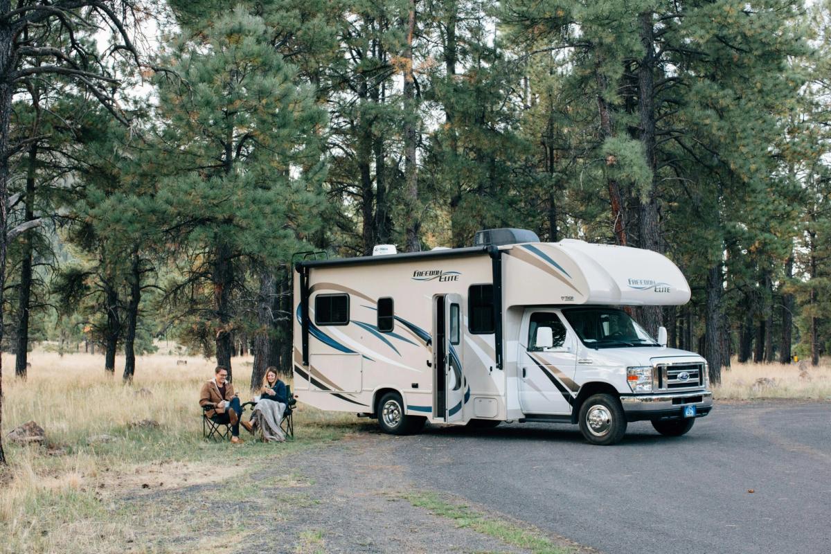Couple relaxing in camping chairs beside a white Freedom Elite motorhome parked among tall pine trees in a forest clearing.