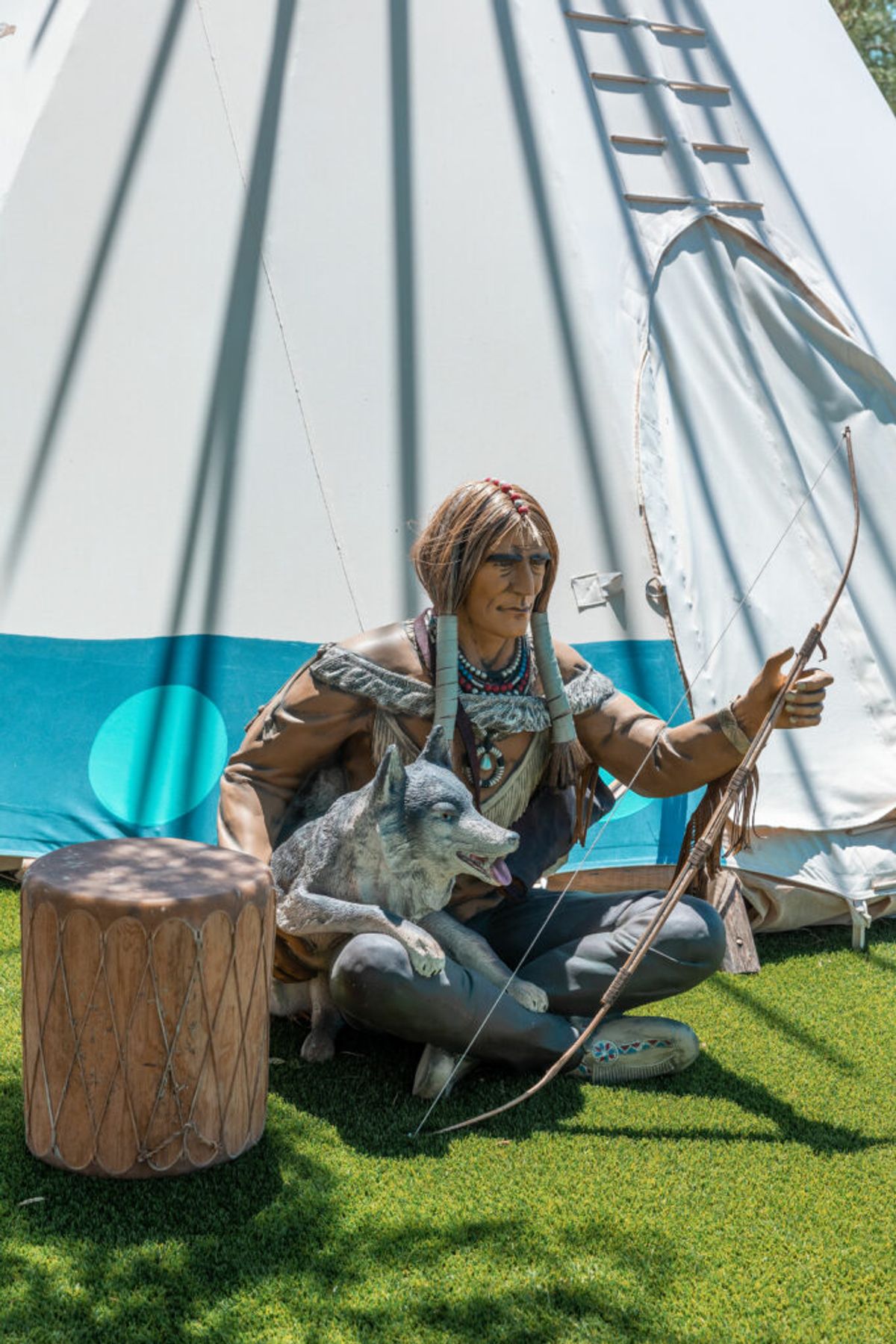 Person in traditional Native American attire sitting cross-legged with bow and arrow next to drum and dog in front of white teepee on grass