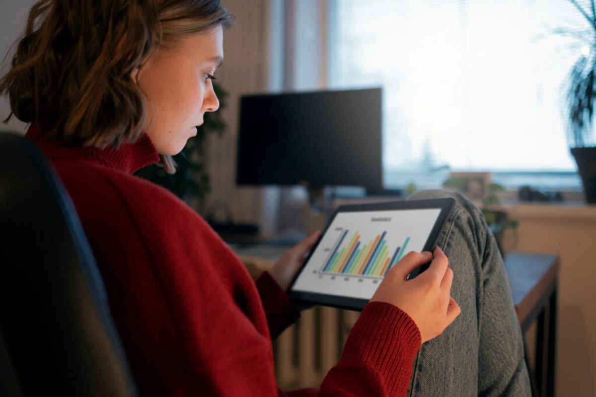 Woman in red sweater viewing bar chart data on tablet while sitting at desk with computer monitor in background.