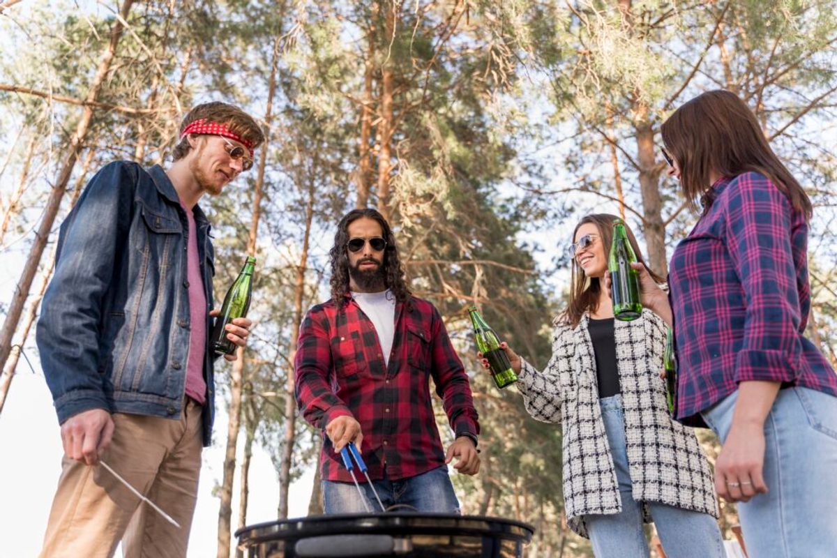 Group of five friends gathered around a fire pit in a forest clearing, holding beer bottles and socializing outdoors.