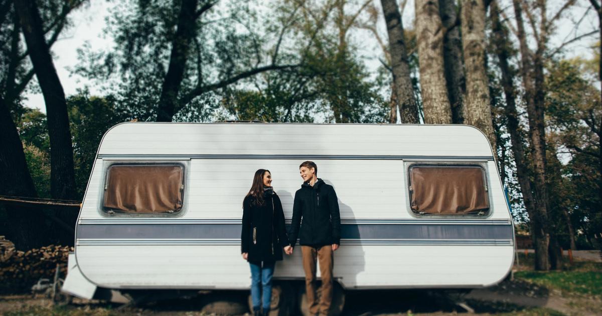 Couple standing outside a white travel trailer in a wooded campground setting, discussing RV park amenities and accommodations.
