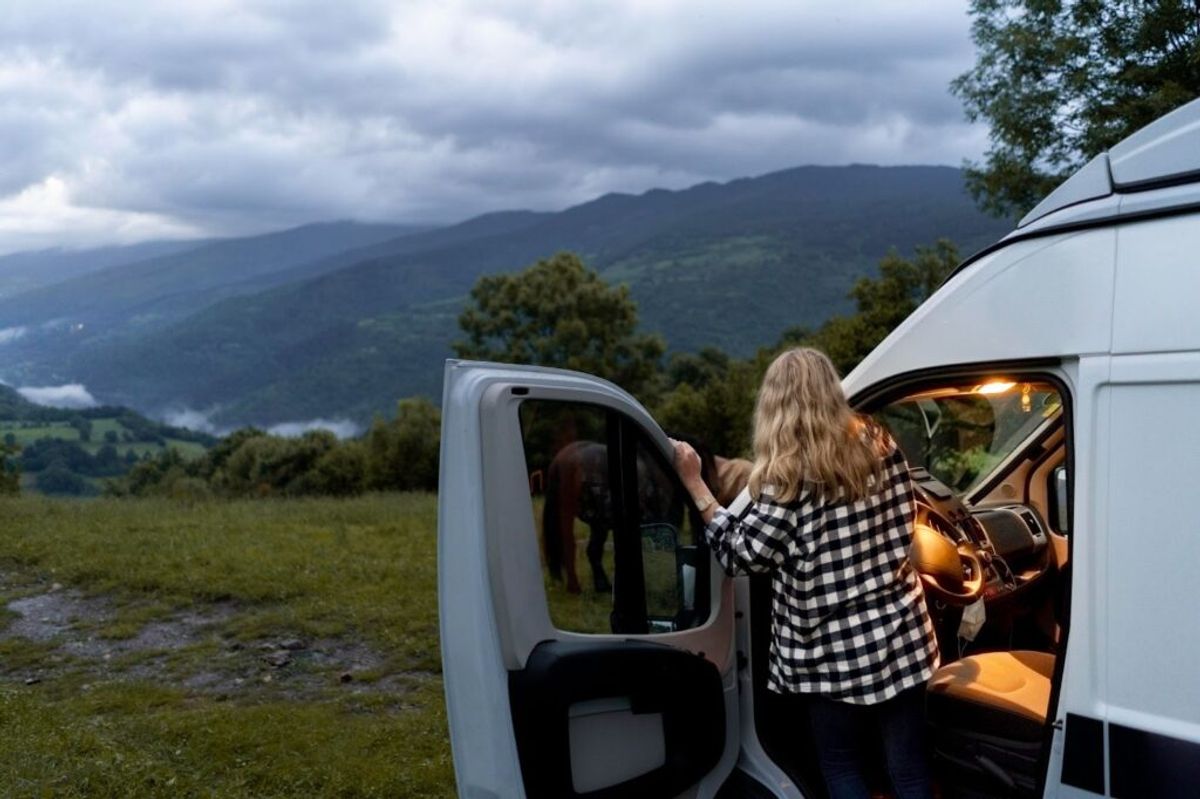 Woman in checkered shirt standing between two white vans in a mountainous valley landscape with trees and cloudy sky.
