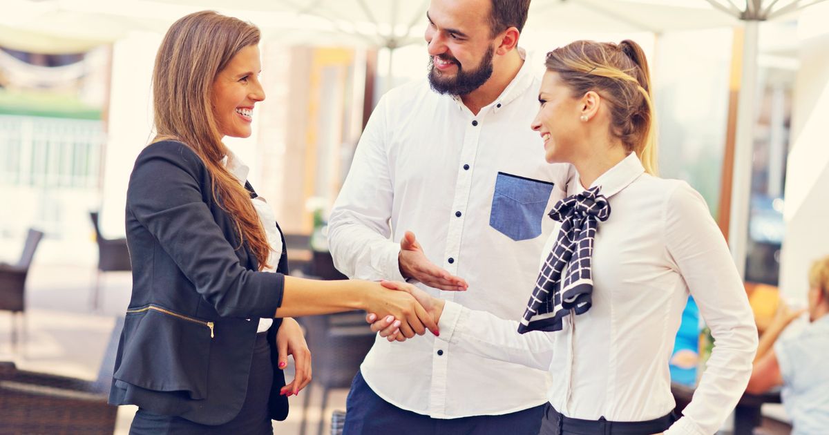 Three business professionals shaking hands and smiling in a modern office setting with umbrellas and glass partitions in the background.
