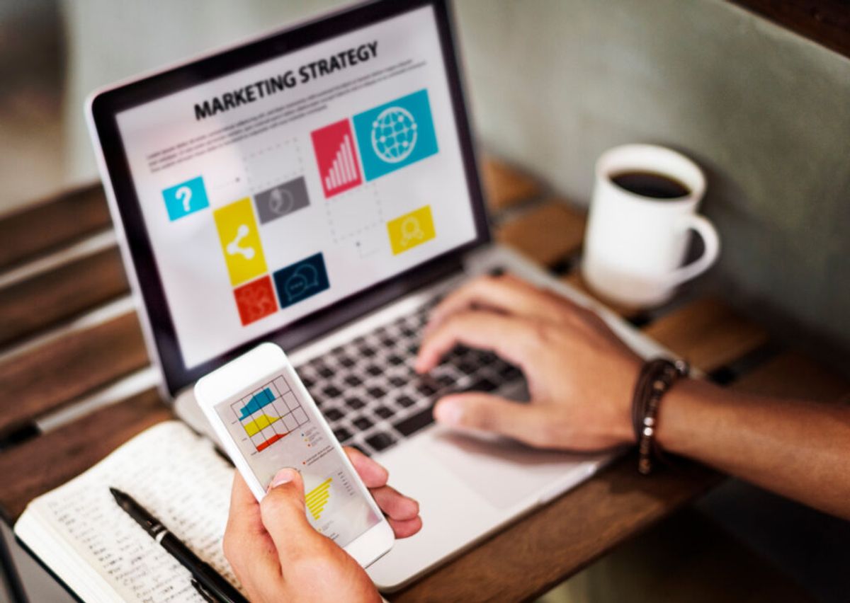 Person holding smartphone displaying marketing strategy charts while working at laptop with coffee cup on wooden desk