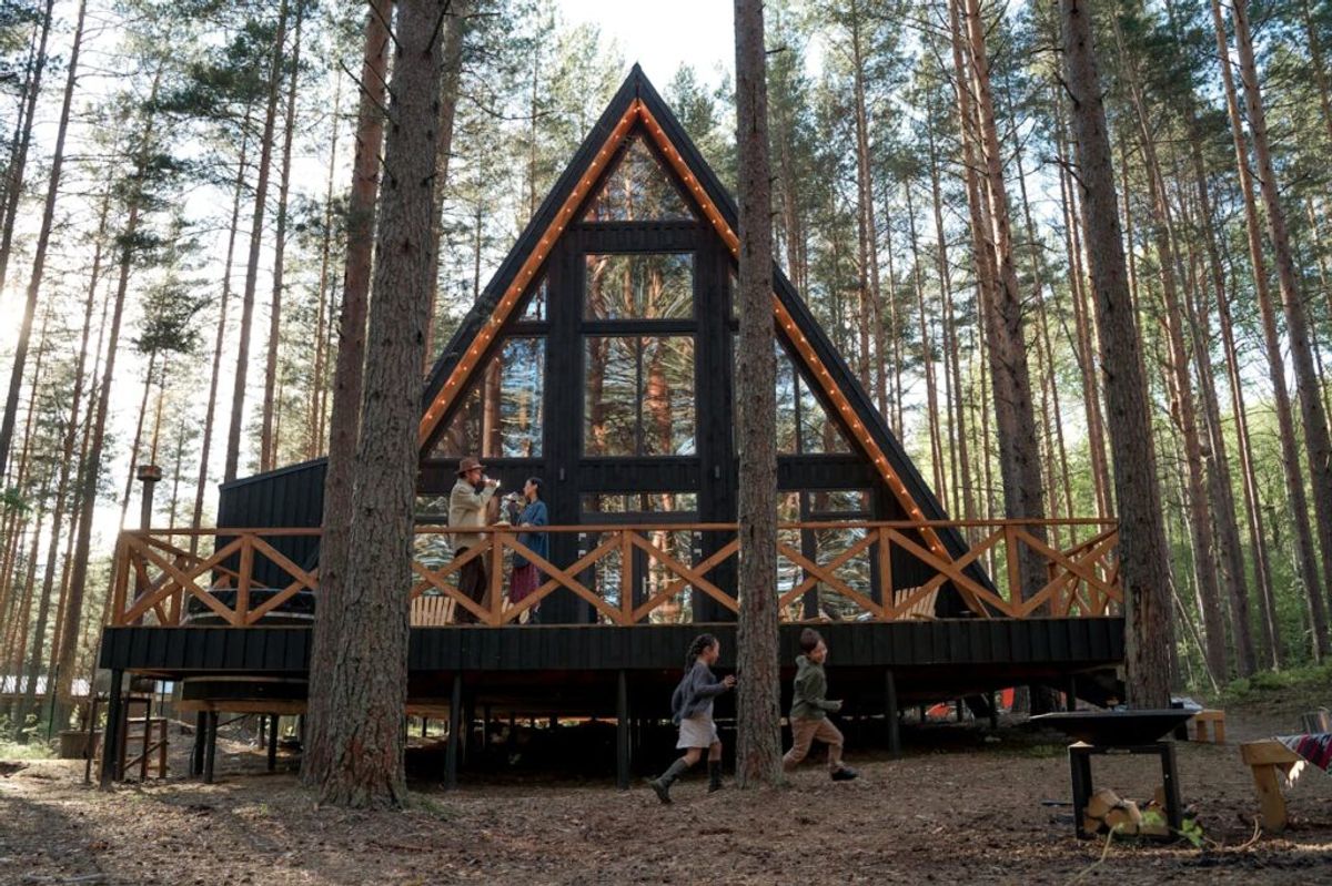 Modern A-frame glamping cabin with large windows nestled among tall pine trees, guests relaxing on wraparound wooden deck.