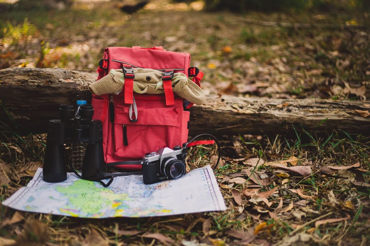 Red backpack with binoculars, camera, and topographic map arranged on forest ground next to fallen log, symbolizing outdoor adventure gear.
