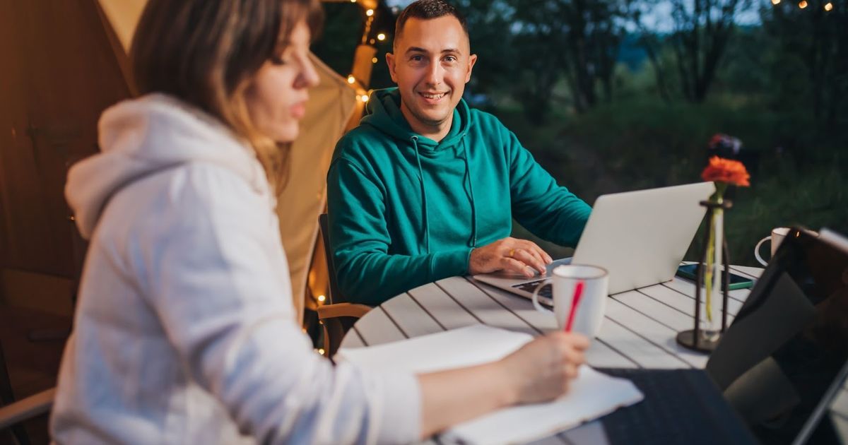 Two people reviewing financial documents at an outdoor table under a yellow awning at dusk, with a laptop and paperwork visible.