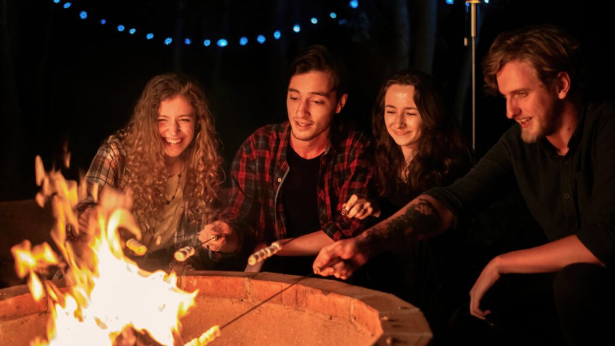 Four young adults roasting marshmallows around a glowing fire pit at night under string lights, enjoying outdoor recreation together.