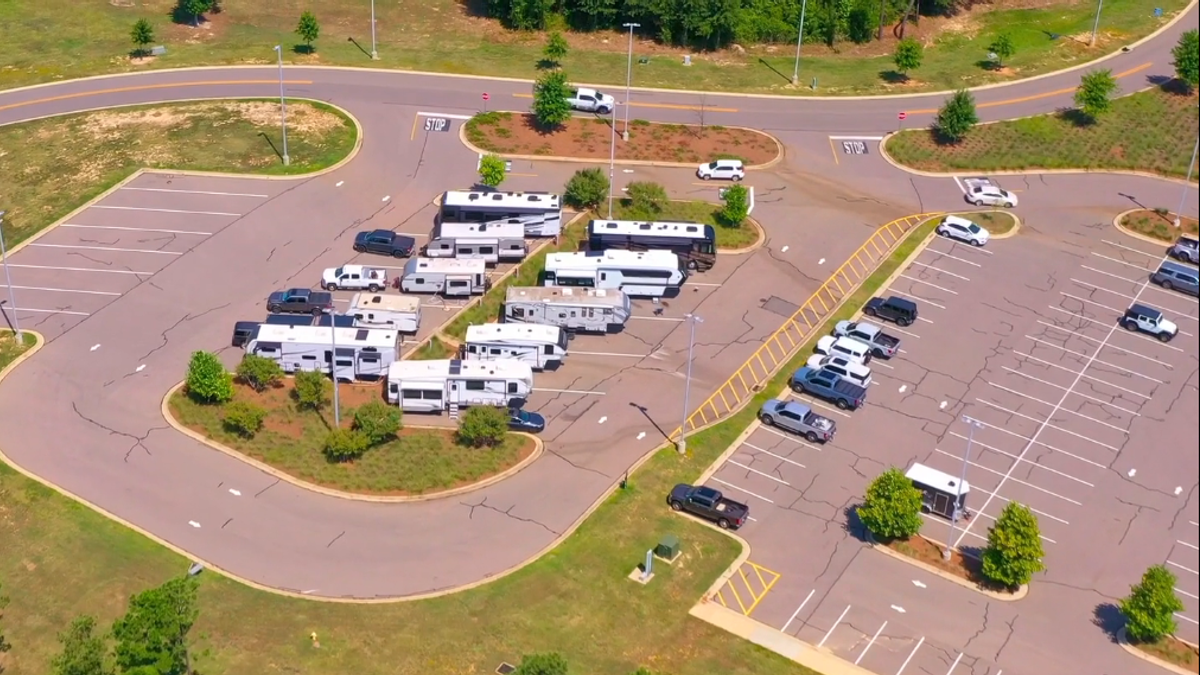 Aerial view of an RV park with multiple white recreational vehicles parked in organized rows, surrounded by landscaping, paved roads, and parking spaces.