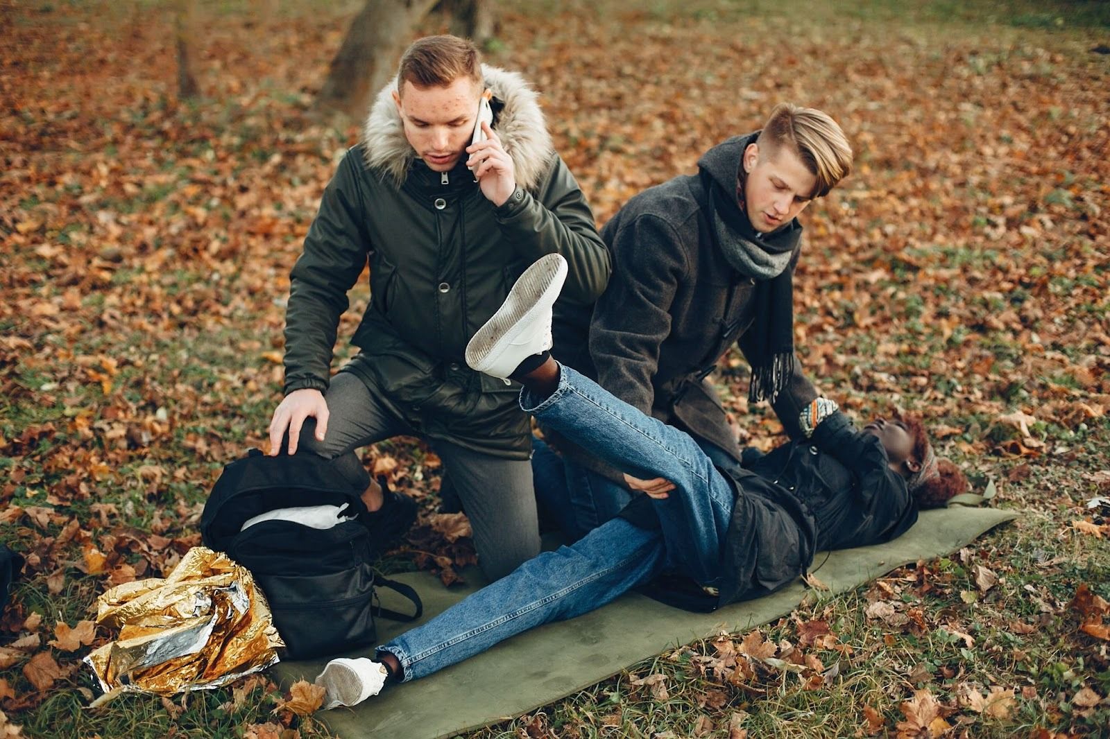 Two people attending to an injured person lying on the ground in a forest, with one person on the phone, depicting an emergency situation.