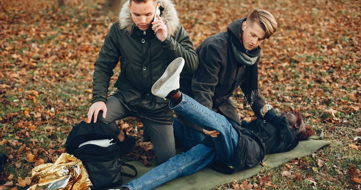 Two people attending to an injured person lying on the ground in a forest, with one person on the phone, depicting an emergency situation.