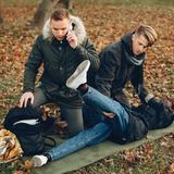 Two people attending to an injured person lying on the ground in a forest, with one person on the phone, depicting an emergency situation.
