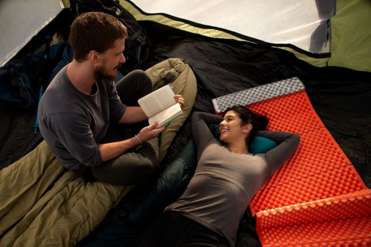 Two campers relaxing inside a tent on sleeping pads, one reading a book while the other listens, surrounded by colorful bedding and gear.