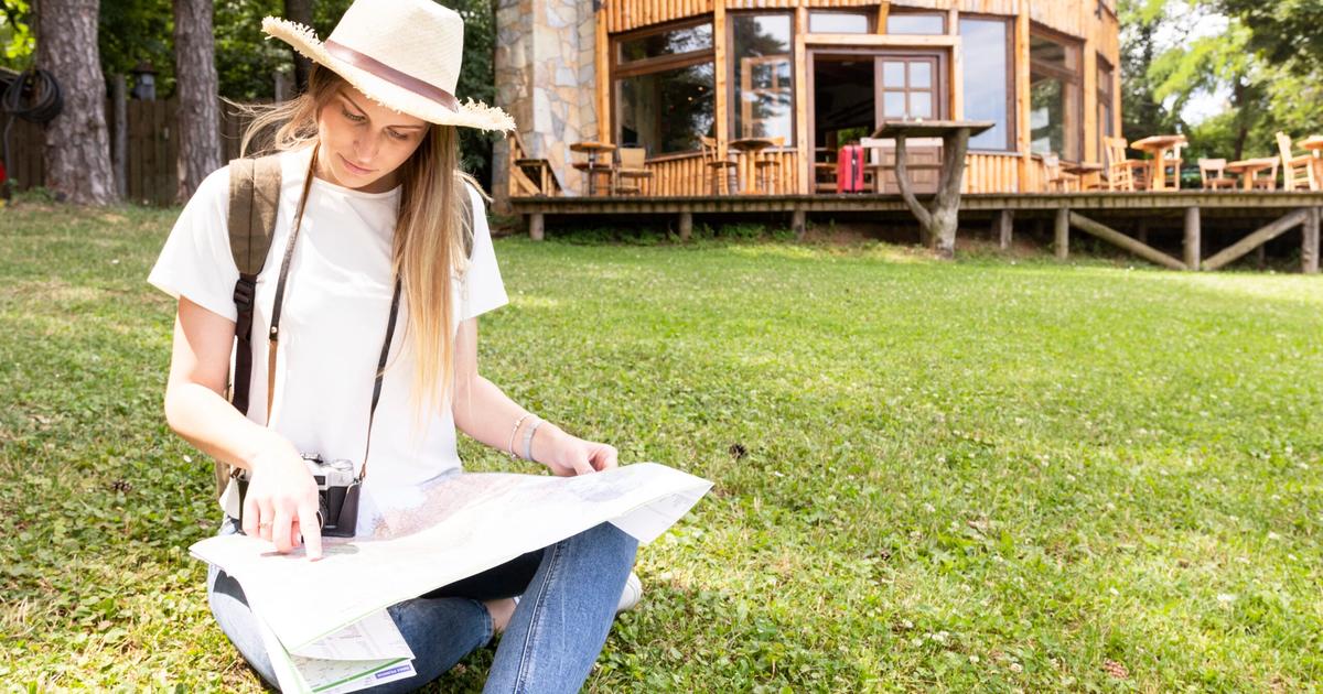 Young woman in white hat studying map while sitting on grass near modern wooden cabin with deck and trees.