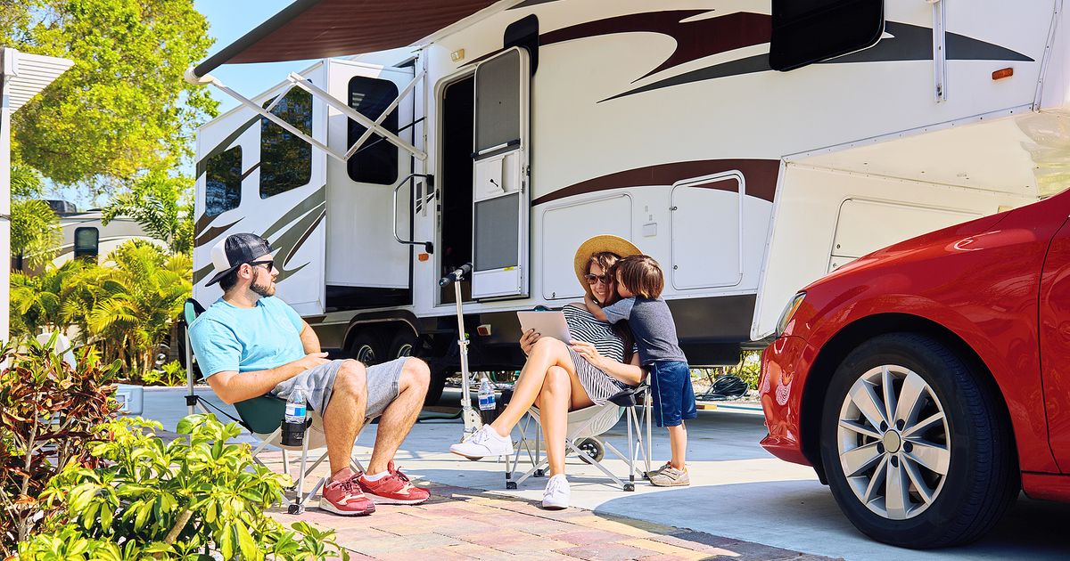 Mother father and son sitting near camping trailer smiling. Woman men kid relaxing on chairs near car and palms. Family spending time together on vacation near sea or ocean in modern rv park