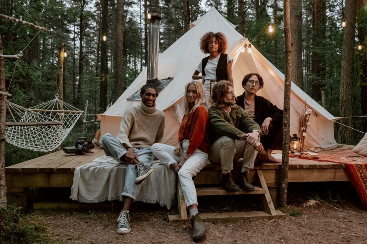 Five young adults relaxing outside a glamorous white bell tent in a forest setting, with hanging lights and hammock visible nearby.