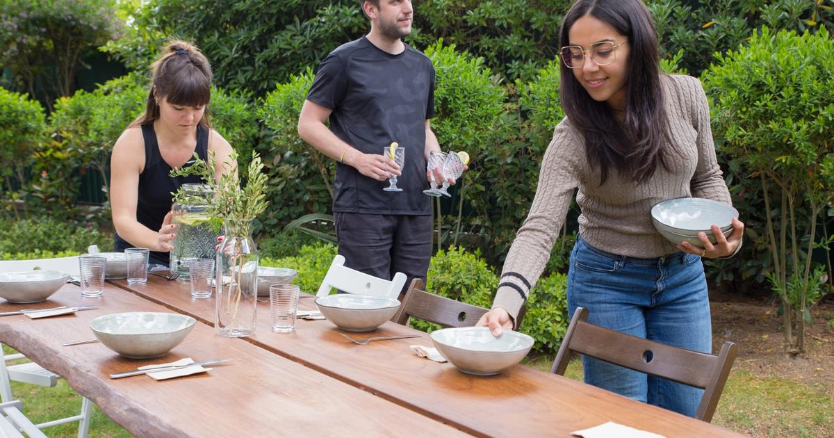 Three people setting an outdoor dining table in a garden with bowls, glasses, and flowers for a meal celebration.
