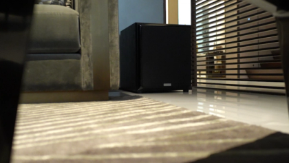 view of the XSCACE Juniper deep-bass subwoofer sitting on rubber isolation feet on a hardwood floor, highlighting its powerful exposed woofer cone engineered for immersive home audio.
