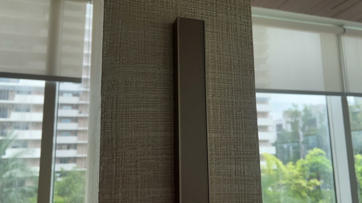 Close-up of a champagne-finish XSCACE QuadCane slim array speaker surface-mounted on a textured plaster pier between floor-to-ceiling gym windows